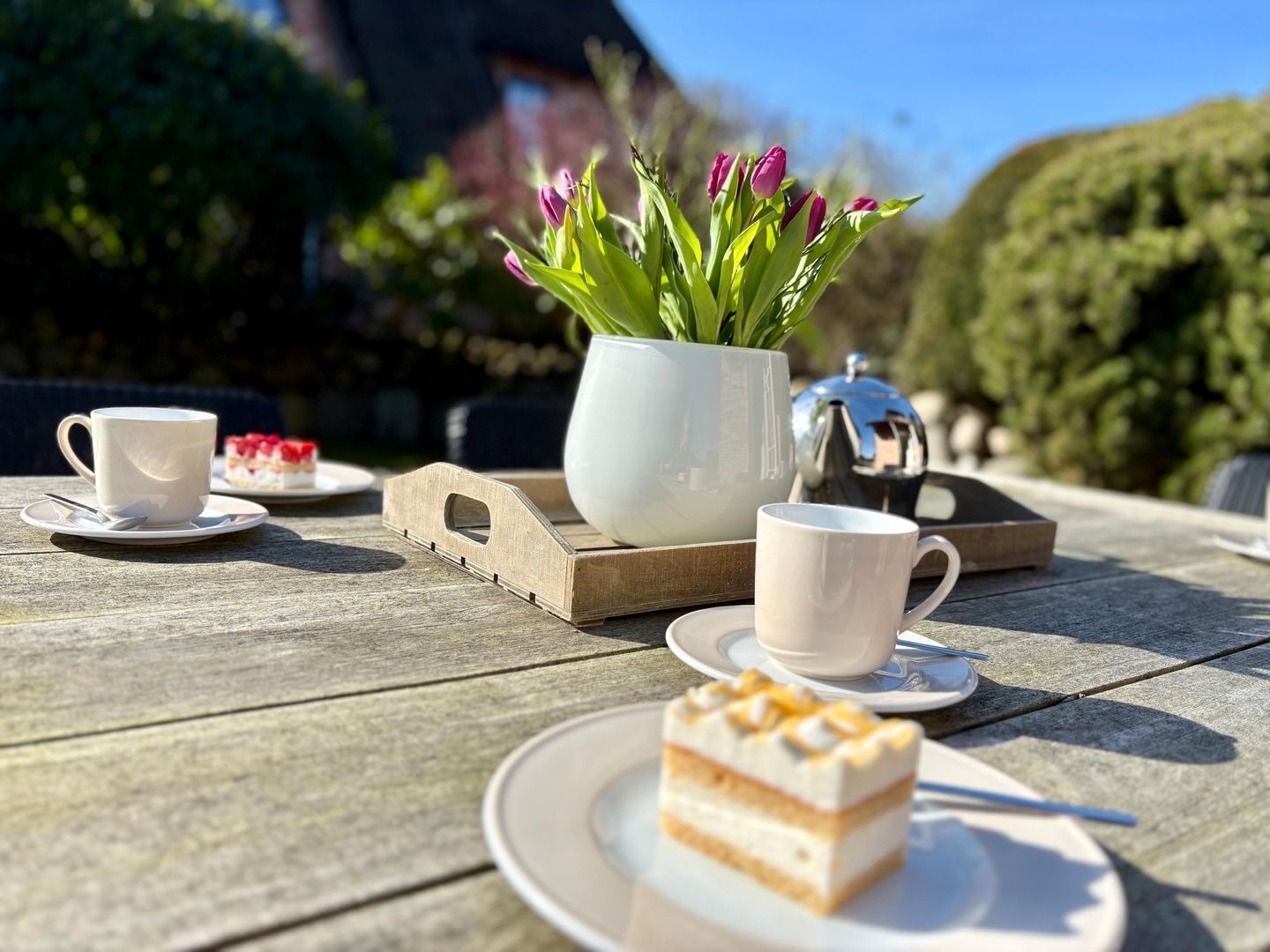 Une table en bois sur laquelle se trouvent une tasse blanche, une soucoupe, une petite assiette, un pot blanc avec des fleurs et une part de gâteau.