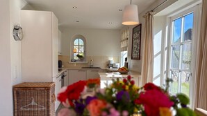 Une cuisine moderne avec des meubles blancs, une table à manger agrémentée d’un vase de fleurs et une grande fenêtre avec des rideaux.