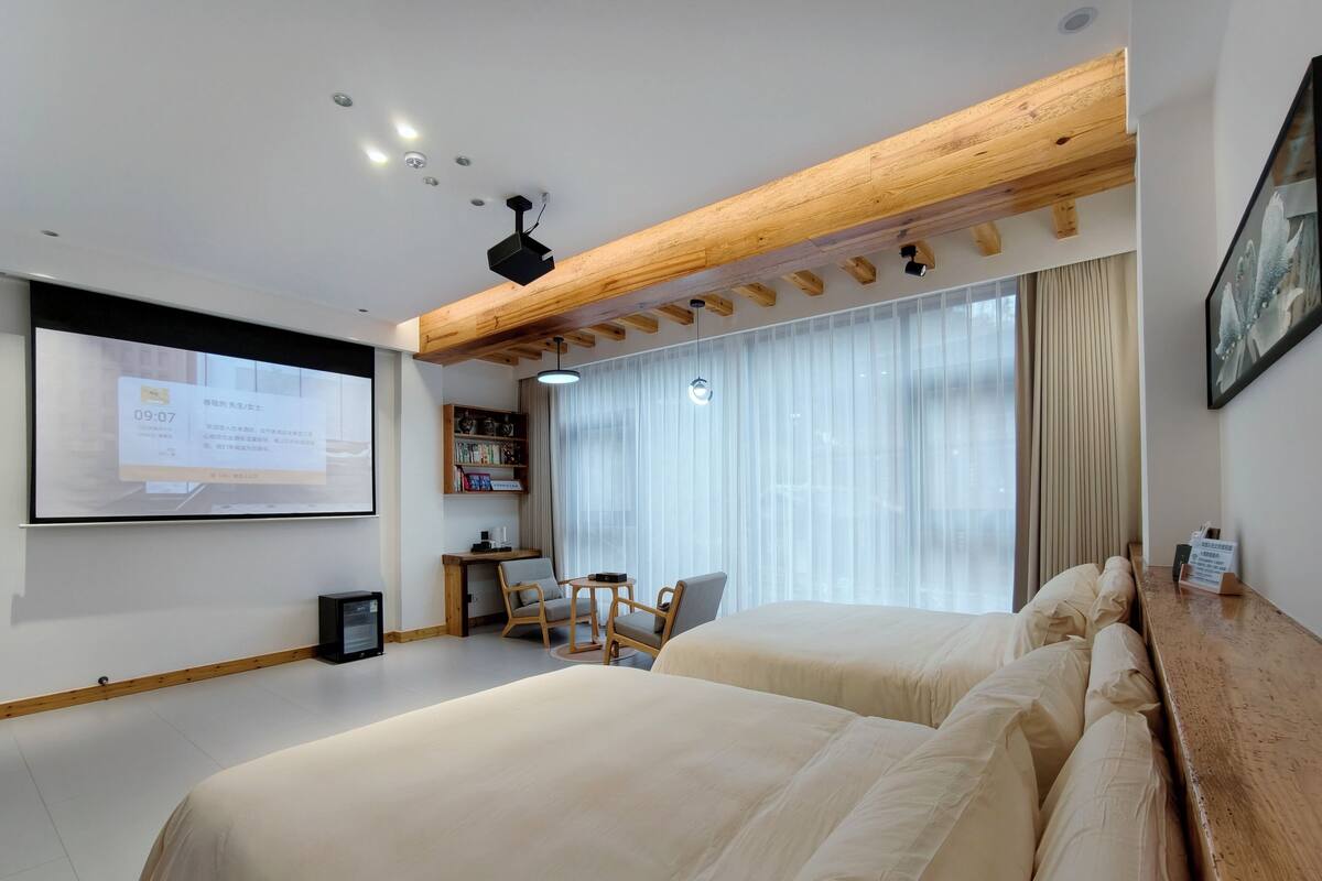 Una habitación de hotel con una cama grande, un escritorio con silla, una estantería y un ventanal con cortinas.