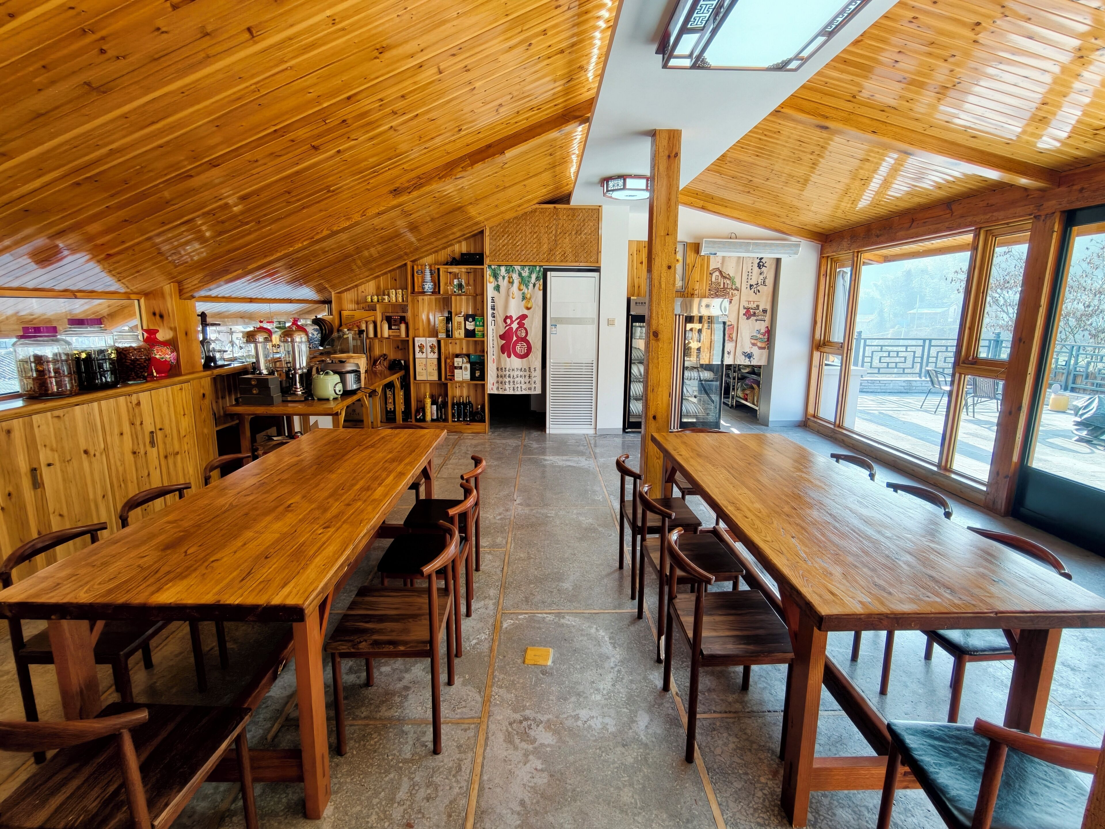 Un vaste espace dédié à la salle à manger, avec des tables et des chaises en bois, un coin cuisine doté d’étagères remplies de bocaux et de bouteilles, et de grandes fenêtres offrant une vue sur l’extérieur.