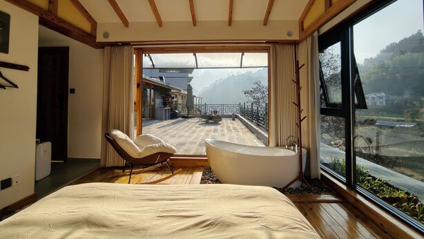 A bedroom with a large bed, a sitting area with a chair, a bathtub, and a view of a mountainous landscape through a large window. - Zhangjiajie Zhiguiju (Zhangjiajie)