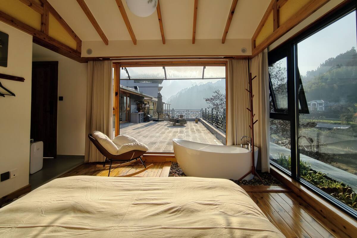 Un dormitorio con una cama grande, una zona de estar con una silla, una bañera y vistas a un paisaje montañoso a través de un ventanal amplio.