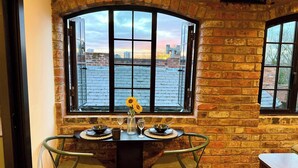 Un comedor con pared de ladrillo, un ventanal con vista a la ciudad, dos sillas, una mesa con un tazón y copas de vino, y un mueble pequeño.