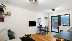 Un saló modern amb un sofà, una televisió de pantalla plana, una zona de menjador amb cadires, una taula petita i una vista per una finestra gran.
