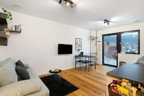 Una sala de estar moderna con un sofá, un televisor de pantalla plana, una zona de comedor con sillas, una mesita y vistas a través de un ventanal grande.