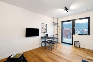Un salon moderne comprenant un coin repas, un téléviseur à écran plat fixé au mur, une fenêtre donnant sur la ville et un tabouret noir.