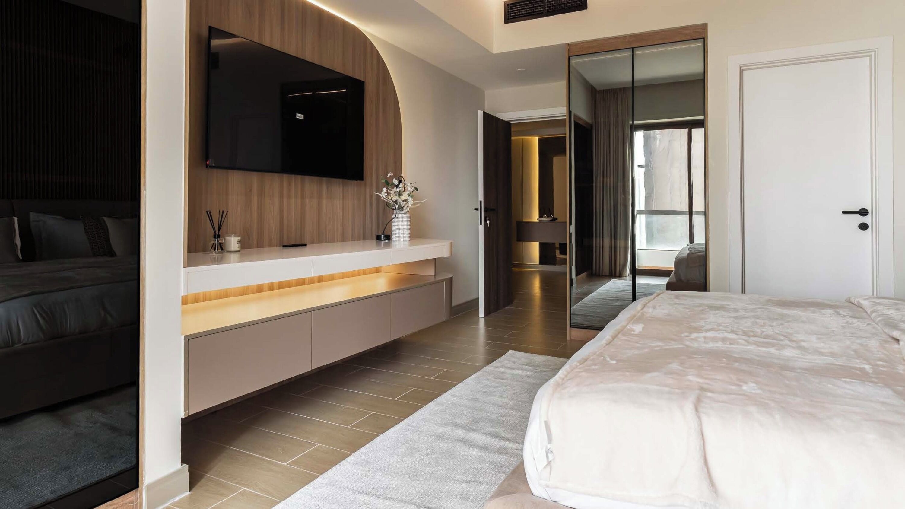A modern bedroom with a large bed, a built-in TV, a bedside table with a vase of flowers, and a sliding door leading to a bathroom.