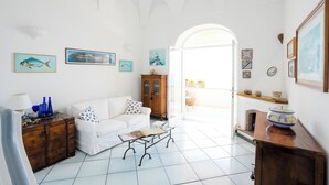 Ein helles Wohnzimmer mit einem weißen Sofa, einem Holzschrank und einem Glastisch. An den Wänden befinden sich Bilderrahmen und ein Blick ins Freie durch einen Bogen.