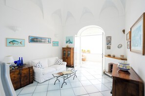 Ein helles Wohnzimmer mit einem weißen Sofa, einem Holzschrank und einem Glastisch. An den Wänden befinden sich Bilderrahmen und ein Blick ins Freie durch einen Bogen.