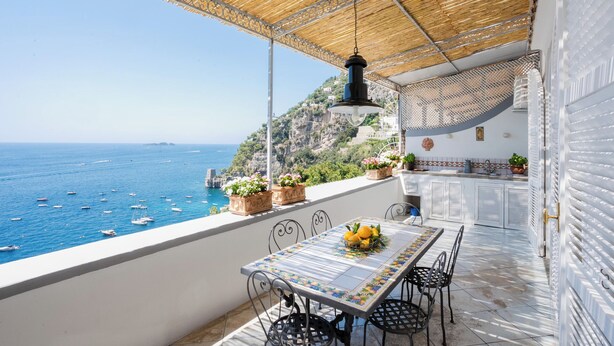 Ein Balkon mit Tisch für zwei Personen, mit Blick auf die Küste, Boote und Klippen.