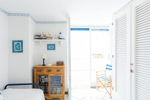 Ein Zimmer mit einem Bett, einem hölzernen Schrank voller Bücher, einem kleinen Tisch mit einer Lampe und einer Tür zu einem Außenbereich mit einem Stuhl.