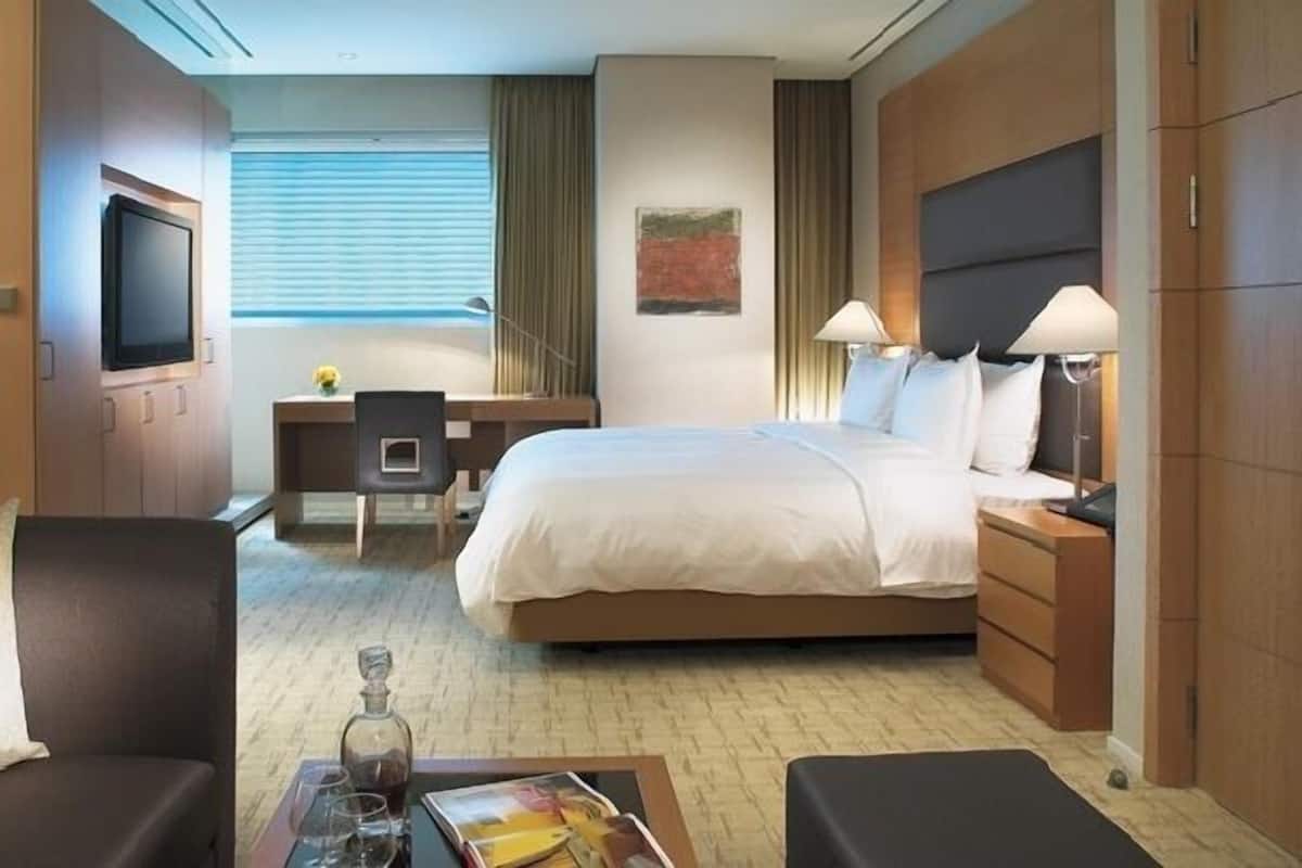 A hotel room with a bed, bedside tables, a desk, a chair, a TV, and a window with blinds.