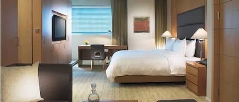 Ein Hotelzimmer mit Bett, Nachttischen, Schreibtisch, Sessel und Fernseher, inklusive Fenster mit Jalousien.