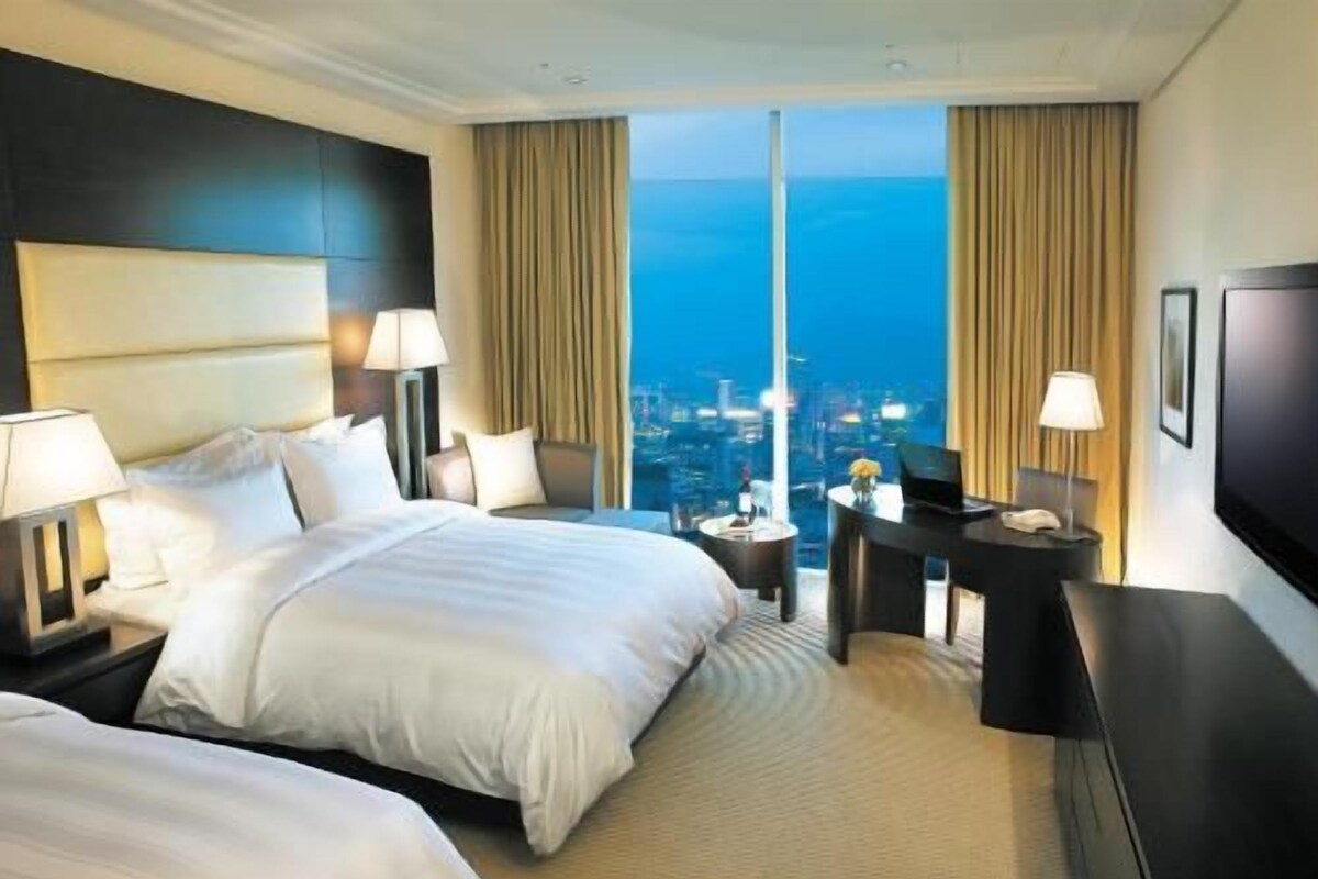 A hotel room with two beds, a desk, a TV, and a city view.