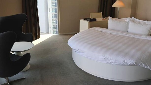 Habitación de hotel con una cama grande, una mesita de centro redonda, una silla moderna, una mesita de noche con lámpara y vista a un edificio a través de la ventana.