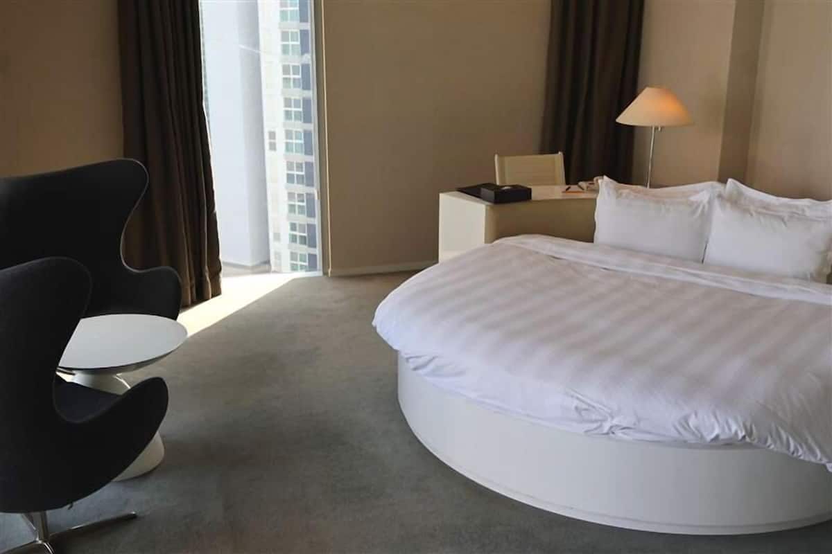 A hotel room with a large bed, a round coffee table, a modern chair, a bedside table with a lamp, and a view of a building through the window.