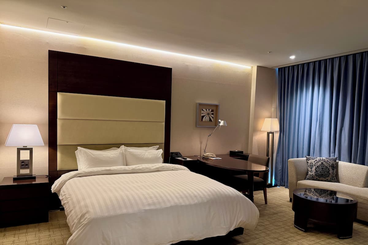 A hotel room with a bed, bedside table, lamp, desk, chair, and a view of the outside through curtains.