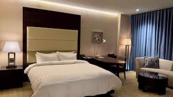 Habitación de hotel con cama, mesita de noche, lámpara, escritorio, silla y vistas al exterior a través de cortinas.