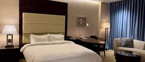 A hotel room with a bed, bedside table, lamp, desk, chair, and a view of the outside through curtains.