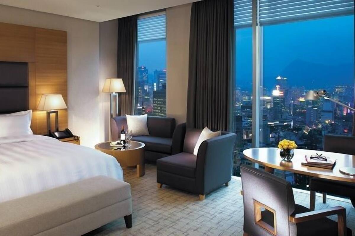 A hotel room with a bed, a desk, a chair, a sofa, and a view of the cityscape.