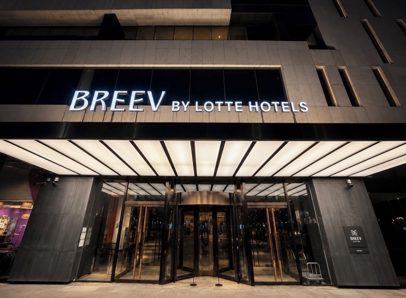 De ingang van BREEV by Lotte Hotels, met een modern design, inclusief een glazen deur en verlichte signage.