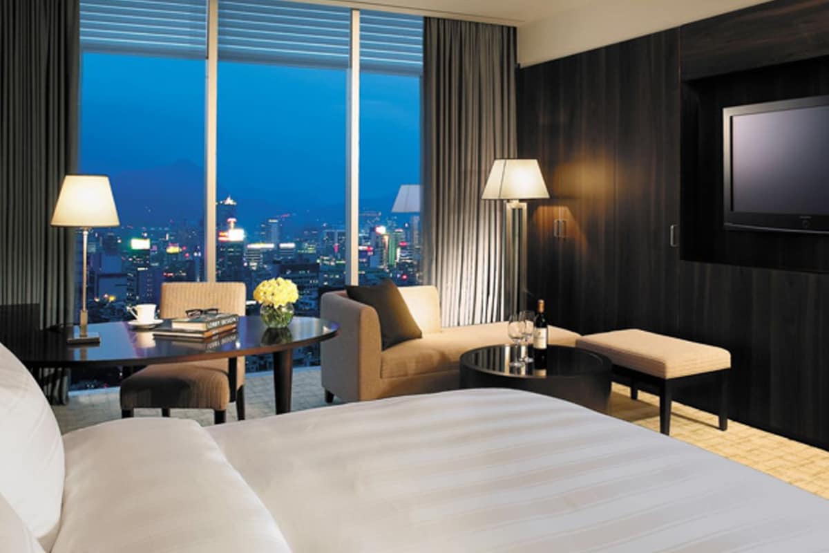 A hotel room with a large bed, a desk with a lamp, a sofa, a small table with a bottle and glasses, and a TV on the wall.