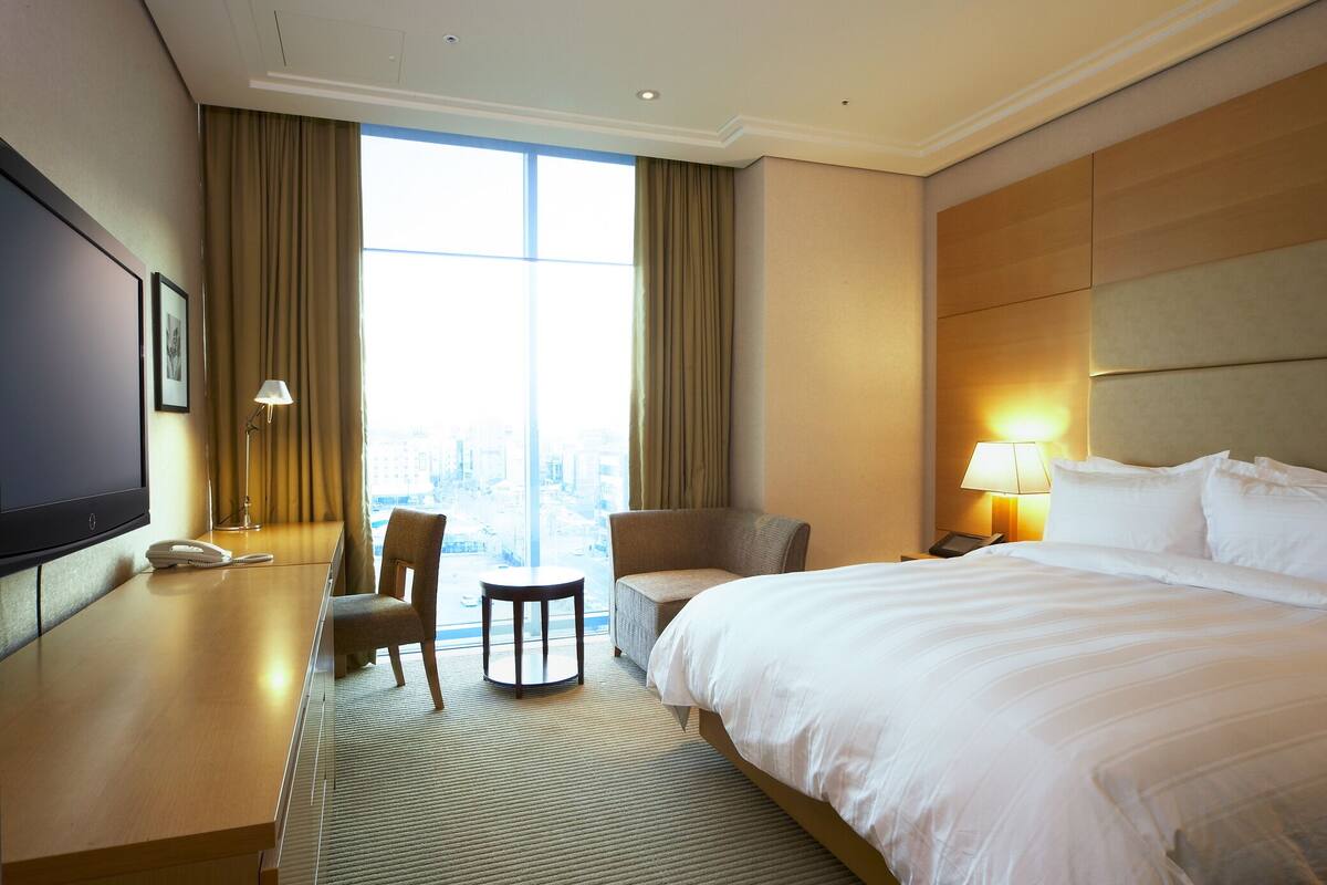 A hotel room with a large bed, a desk, a chair, a small table, a TV, and a window with curtains.