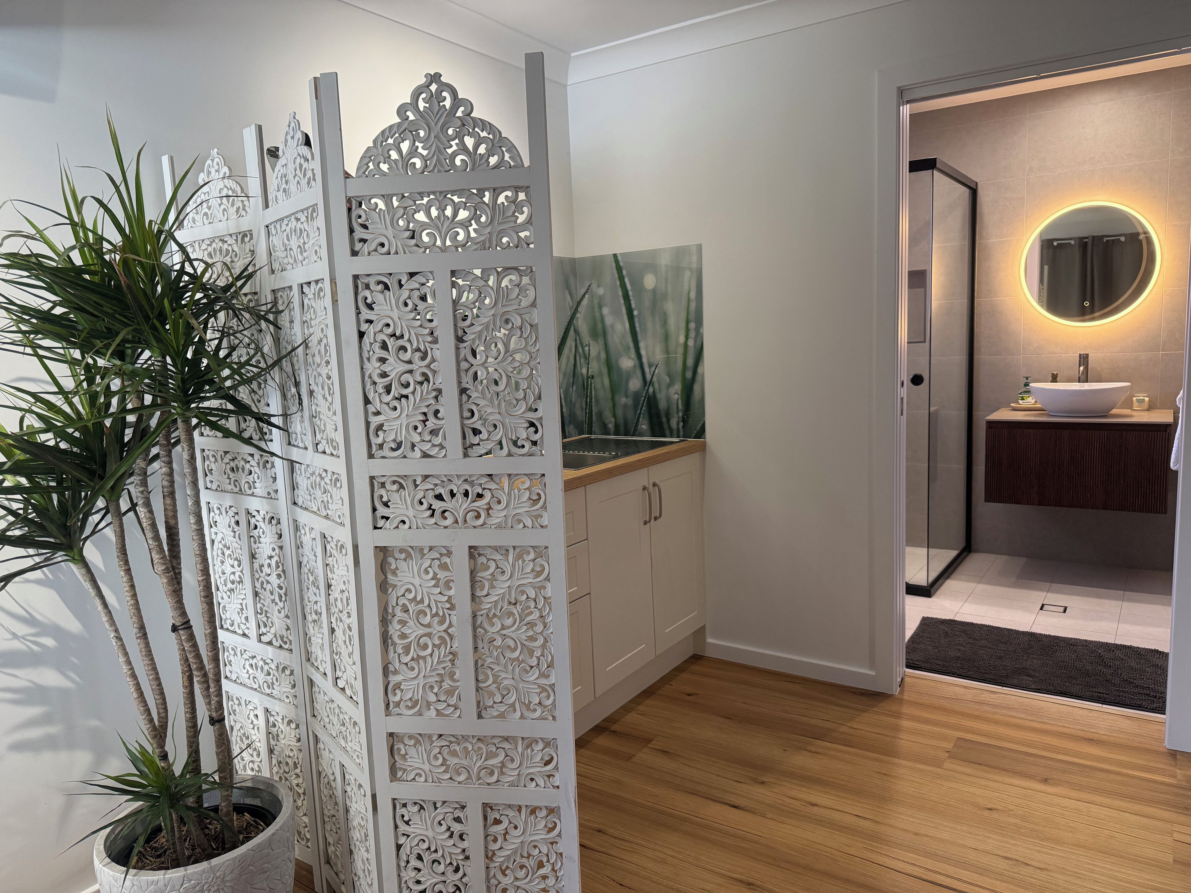 Une salle de bain moderne avec un plancher en bois, un séparateur décoratif blanc et un miroir rond avec un éclairage intégré.