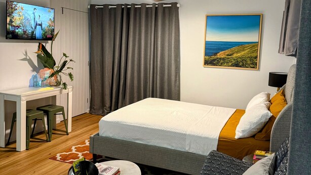 A hotel room with a bed, a desk, a TV, a ceiling fan, and a painting of a landscape. - Town-to-beaches Living I Studio for travelling solo, with bestie, or as couple. (North Boambee Valley)
