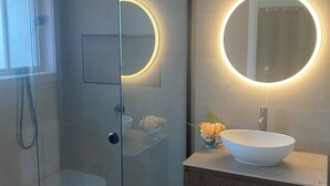 A modern bathroom with a glass-enclosed shower, a round mirror with a light, and a white sink with a decorative bowl on the vanity. - Town-to-beaches Living I Studio for travelling solo, with bestie, or as couple. (North Boambee Valley)