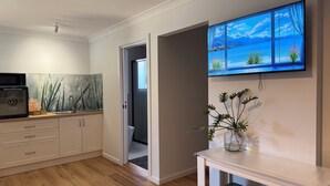 A modern kitchen with wooden flooring, a microwave, a sink, and cabinets. There's a TV mounted on the wall displaying a scenic view. - Town-to-beaches Living I Studio for travelling solo, with bestie, or as couple. (North Boambee Valley)