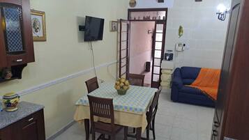 A small dining area with a table set for four, a television mounted on the wall, and a blue sofa in the background.