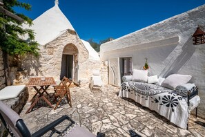 Ein Steinhof mit einer weißen Kuppelkonstruktion, einem Bett mit gemusterten Textilien und einem Tisch mit Stühlen.