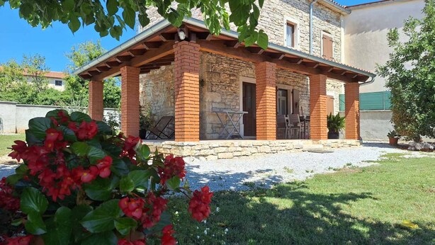 Un edificio in pietra con un portico coperto, una strada di accesso sterrata e un giardino con fiori rossi.