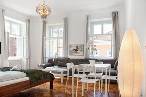 A modern hotel room with a bed, a sofa, a dining table, and chairs. There are windows with curtains, a chandelier, and a potted plant.