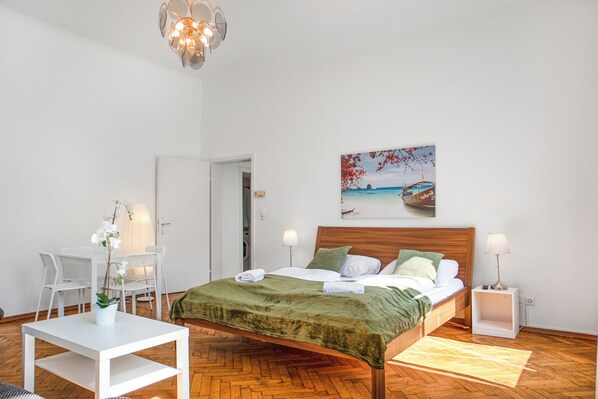 A bedroom with a wooden bed, white walls, a painting of a beach scene, a white desk with a plant, and a wooden floor.
