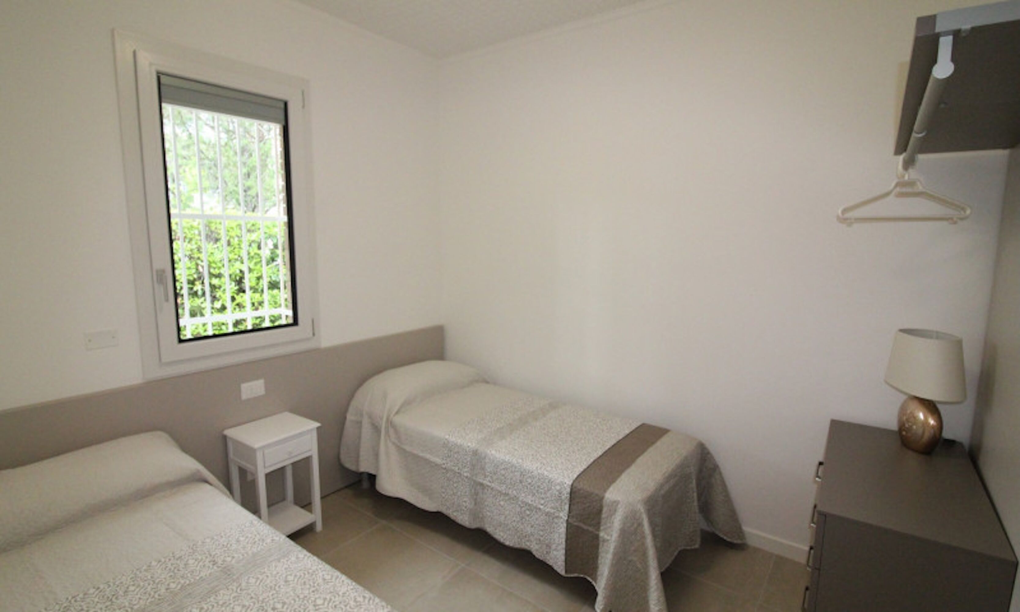 Une chambre simple et épurée, avec deux lits, une petite table de chevet, une fenêtre donnant sur un espace verdoyant et une lampe sur une commode.