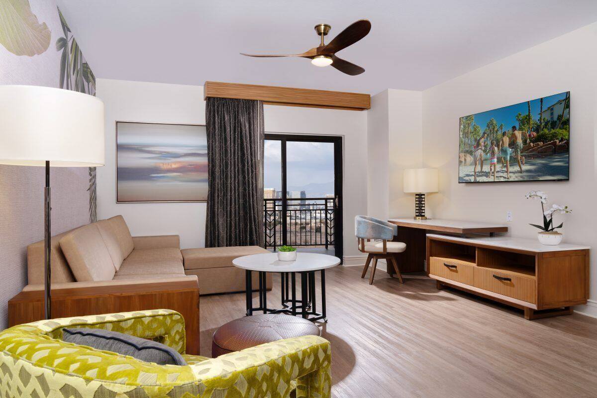 A modern hotel room with a sofa, armchair, dining table, and a ceiling fan.