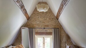 A bedroom with a stone ceiling, a bed with white and striped pillows, a bedside table with a lamp, and a view of the outside through a window with curtains. - The Sun Inn (Bedford)