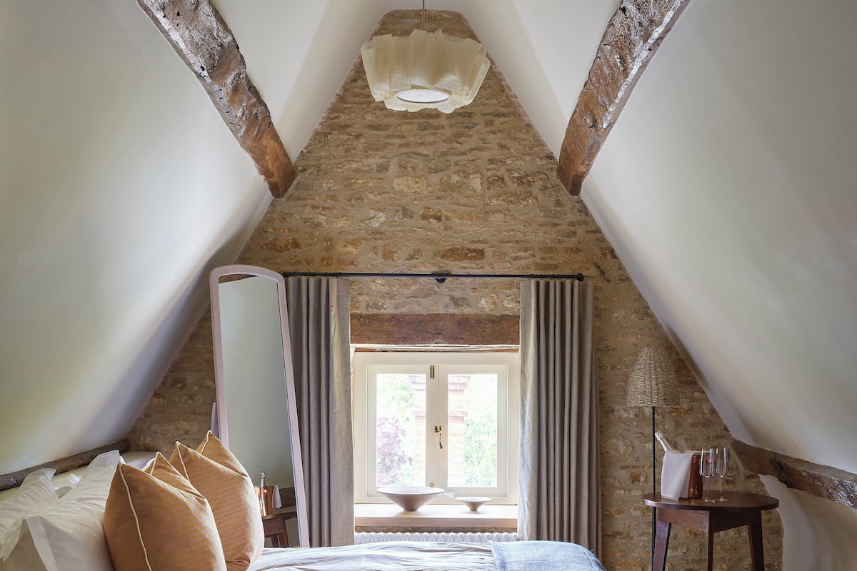 Un dormitorio con techo de piedra, cama con almohadas blancas y a rayas, mesita de noche con lámpara y vistas al exterior a través de una ventana con cortinas.