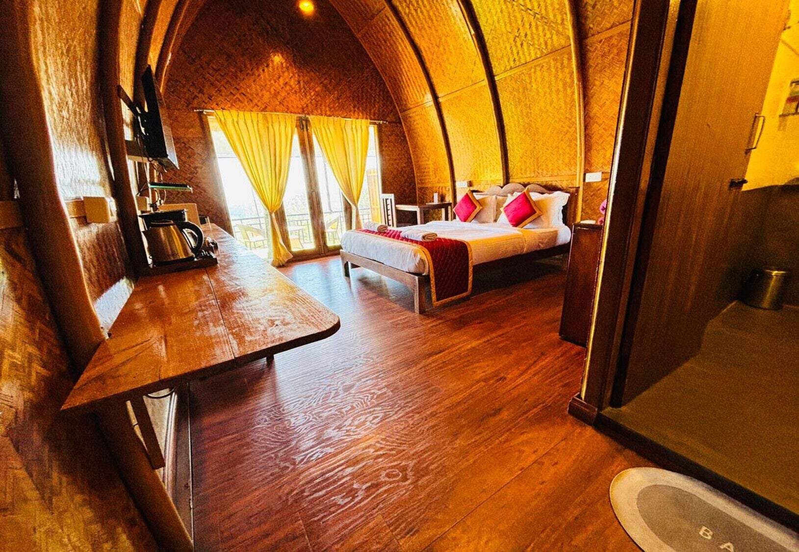 Una habitación acogedora con una mesa de madera, una cama con ropa de cama roja y blanca, y un amplio ventanal con cortinas.