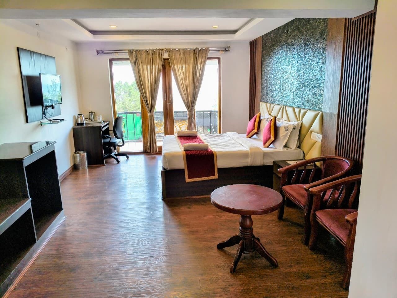 Habitación de hotel con cama, escritorio, silla, televisor y balcón con cortinas.
