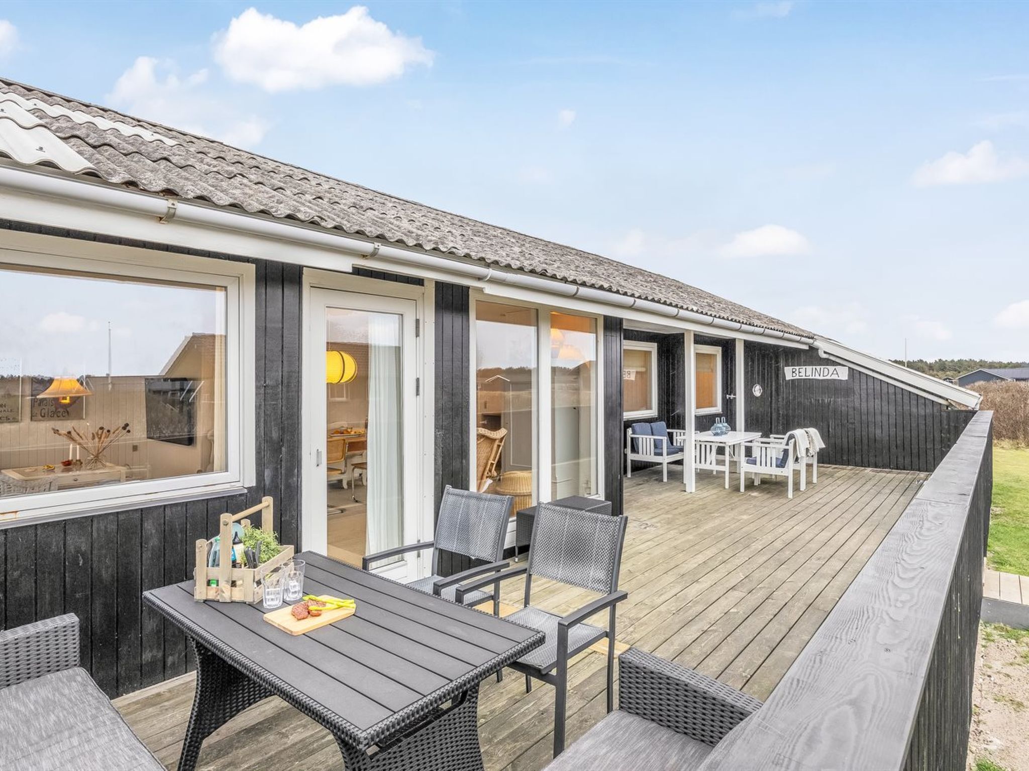 Une terrasse en bois avec des sièges extérieurs et une vue sur l’intérieur d’un bâtiment portant l’inscription « BELINDA ».