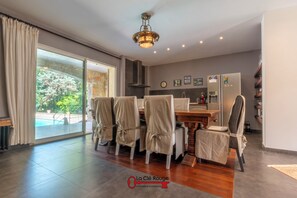 Uma área de jantar com mesa e cadeiras, um lustre, vista para o exterior e uma cozinha com geladeira.