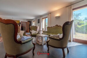 Uma sala de estar ampla com um sofá verde, duas poltronas, uma mesa pequena e vista para o exterior através de grandes janelas.