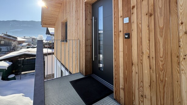 Une façade en bois avec une porte noire, une rambarde en métal et un paillasson noir à l’entrée.