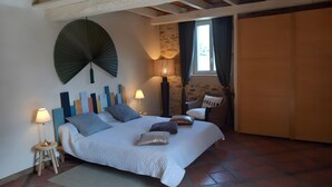 Een slaapkamer met een bed, twee nachtkastjes, een ventilator aan de muur, een raam met gordijnen en een stoel.