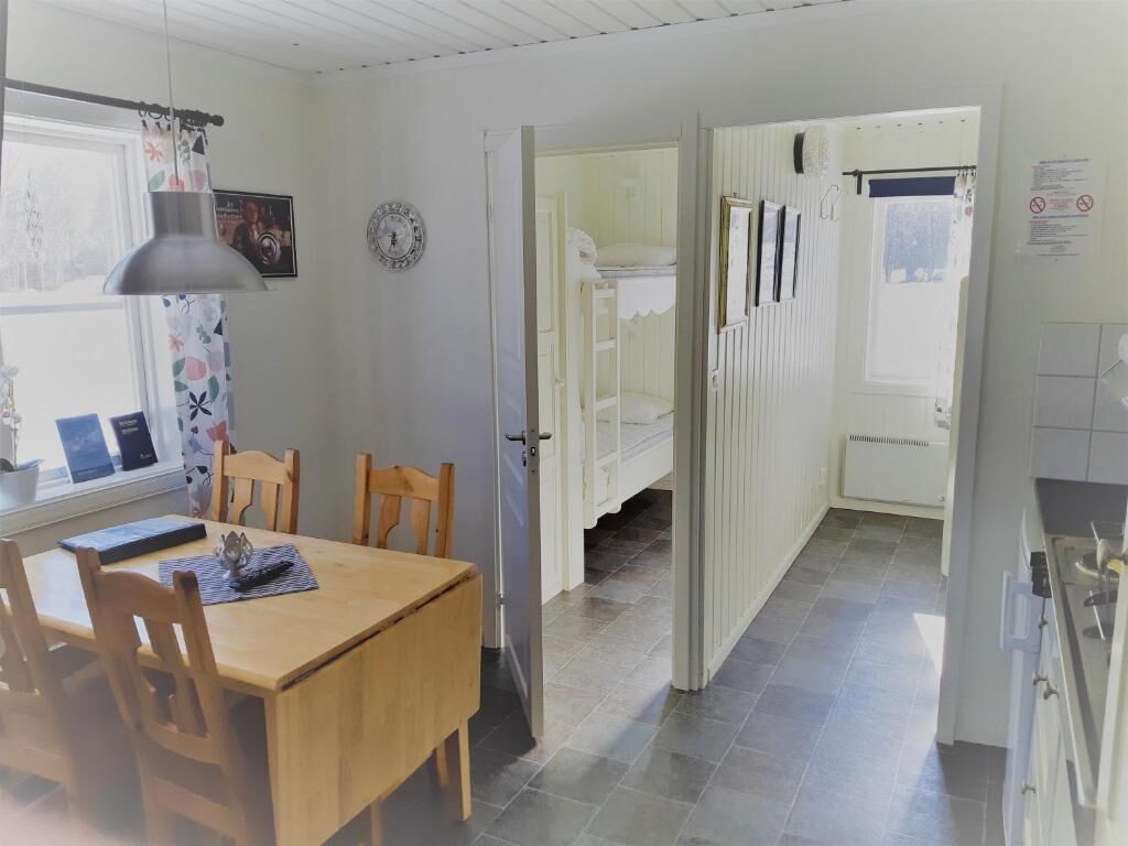 A room with a wooden dining table and chairs, a window with curtains, a door leading to a bathroom, and a kitchen area with cabinets and a stove.