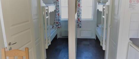 A narrow hallway with bunk beds and a wooden chair, leading to a room with a window and a door.