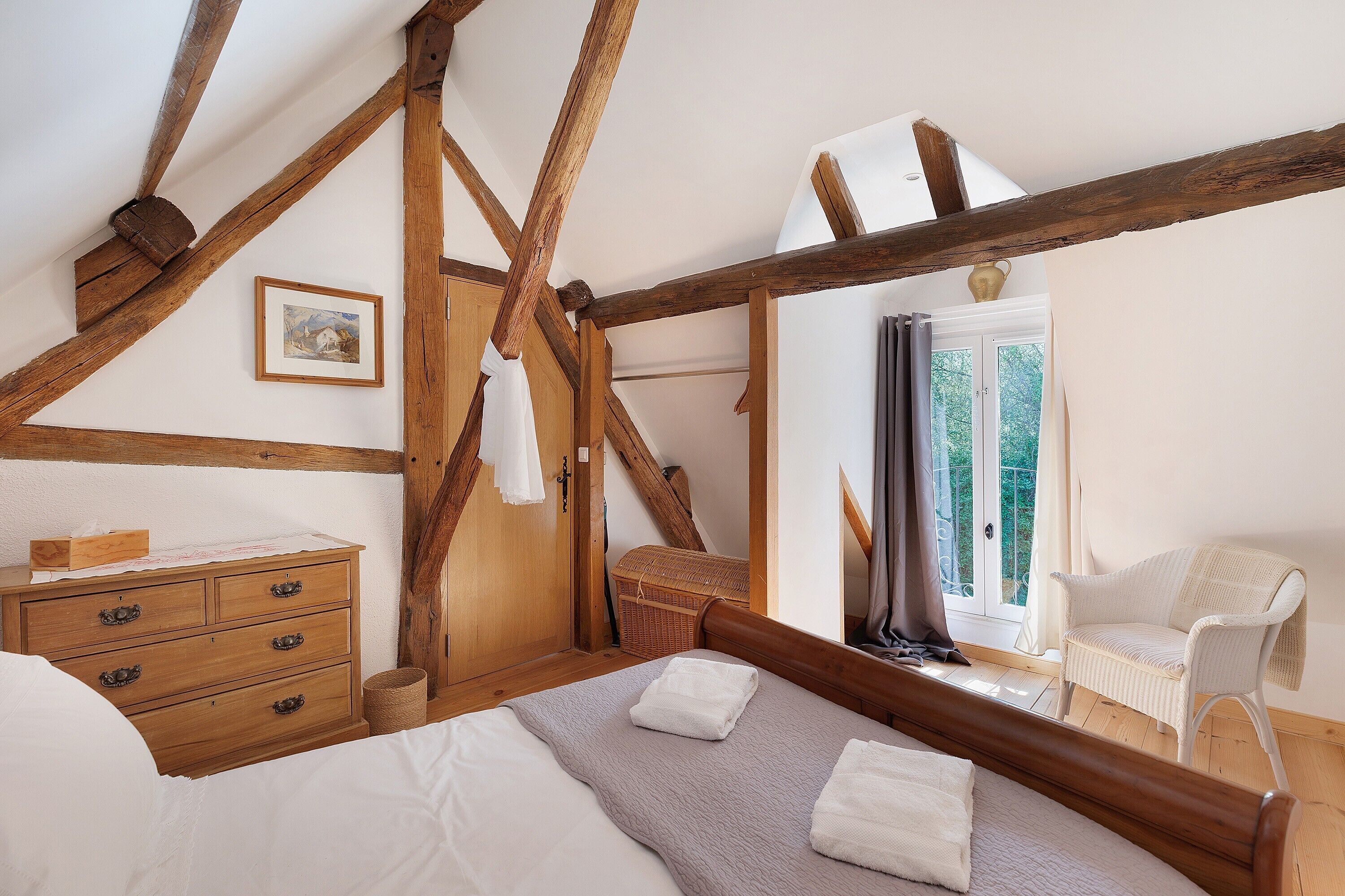A bedroom with a wooden ceiling, a bed with white linens, a dresser, a wicker chair, and a window with curtains.